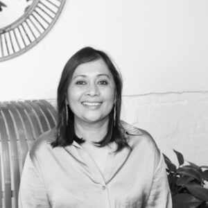 Black and white photo of Pragya Agarwal sitting on a sofa, smiling.