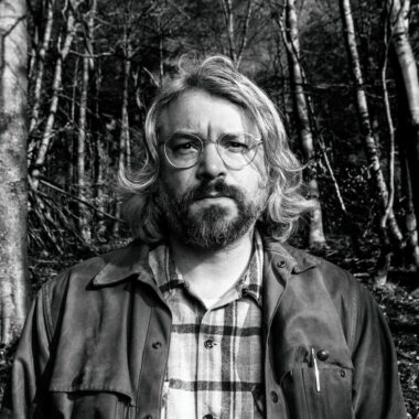 Black and white photo of Will Burns looking at camera and standing in forest